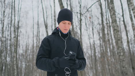 A-young-man-in-a-black-hammer-in-the-winter-is-preparing-to-jog-in-the-park-includes-an-application-for-running-and-runs-with-a-voice-assistant-through-the-forest-doing-business-and-leading-a-healthy-lifestyle.